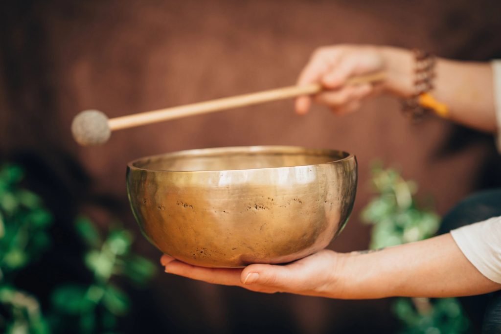 A woman holds a Tibetan healing bowl in her hand. With the other hand, she holds a mallet, poised to strike the bowl. 