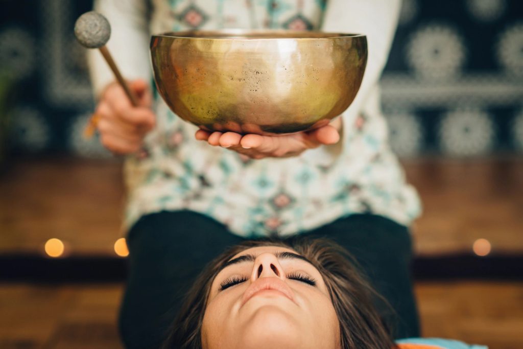 A woman lies on her back with her eyes closed. Behind her, another woman sits with a Tibetan singing bowl in her hand above the woman's head. 