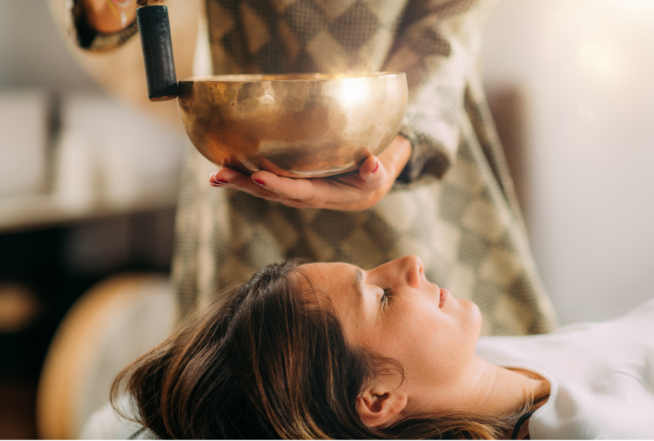 A woman is holding a healing Tibetan bowl over the head of a client, she is vibrating the bowl so the the frequency floats over the client, relaxing them.