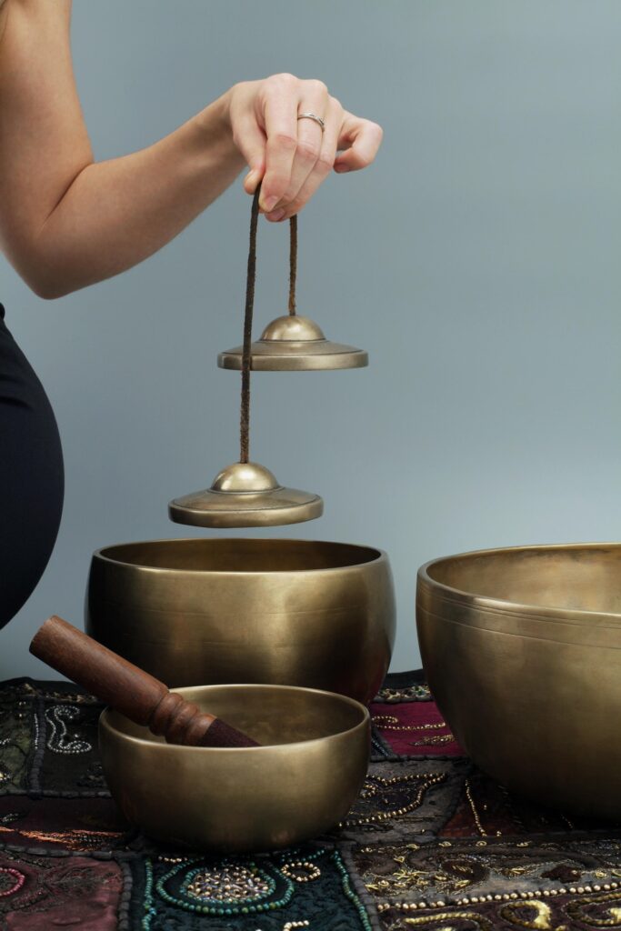 A woman dangles Tingsha bells over three Tibetan bowls sitting on the floor.