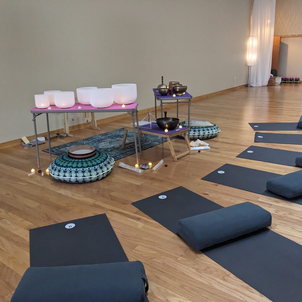 An image of a sound bath journey with sound healing therapy with Lara. Yoga mats are lined up on the floor in front of the sound healing instruments of Tibetan bowls, crystal bowls, and other instruments.