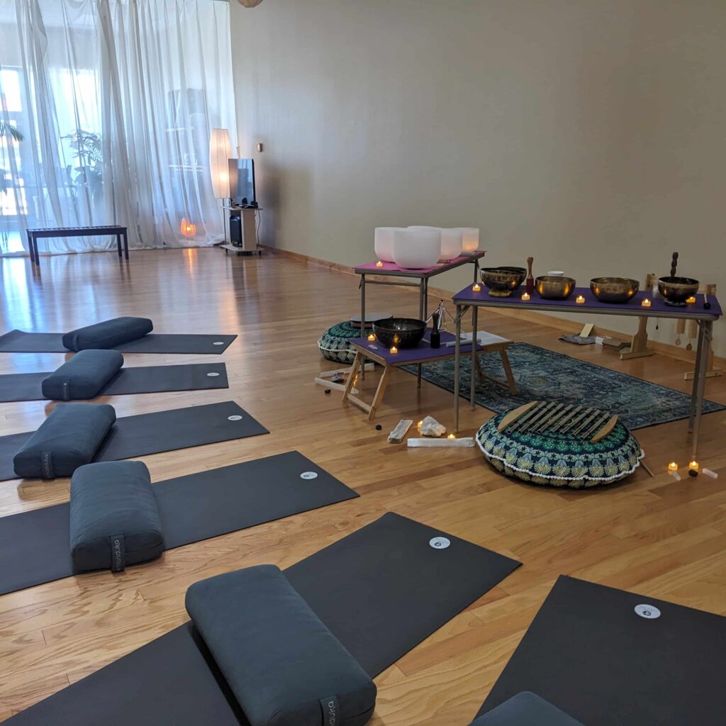 A row of yoga mats are placed in front of sound healing tables with crystal bowls, Tibetan bowls, chimes and other musical instruments.