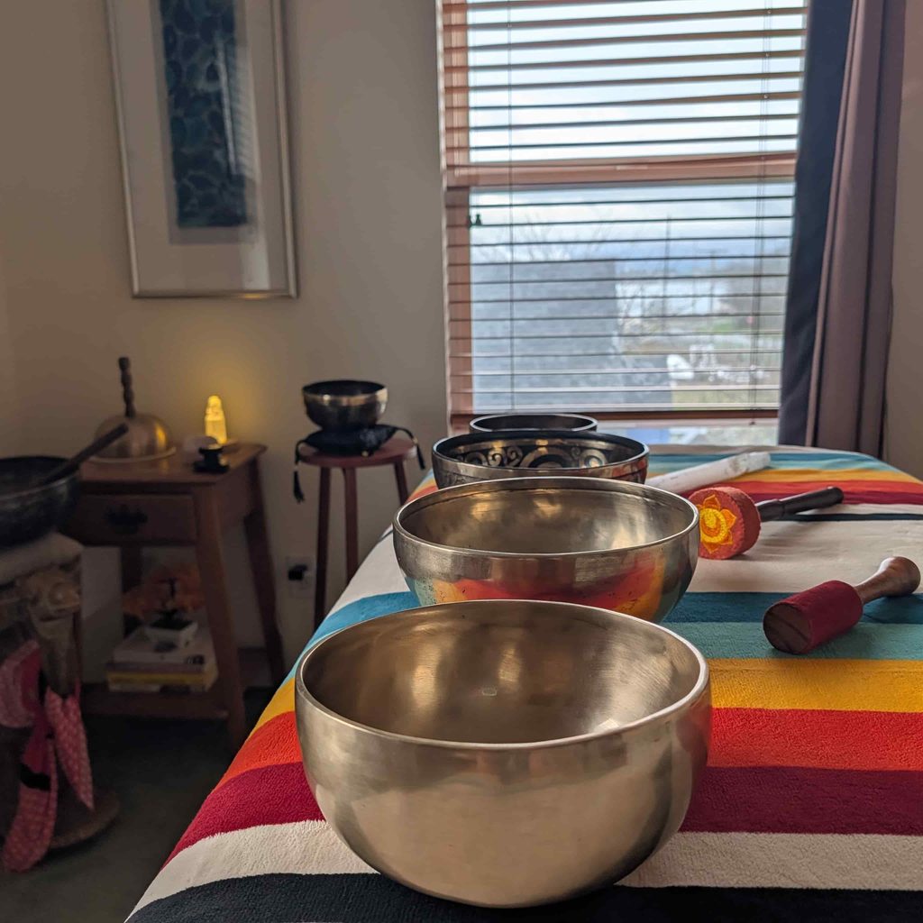Tibetan singing bowls of different sizes lined up on a colorful striped blanket on Lara’s sound healing table, with mallets and a small candle in the background.