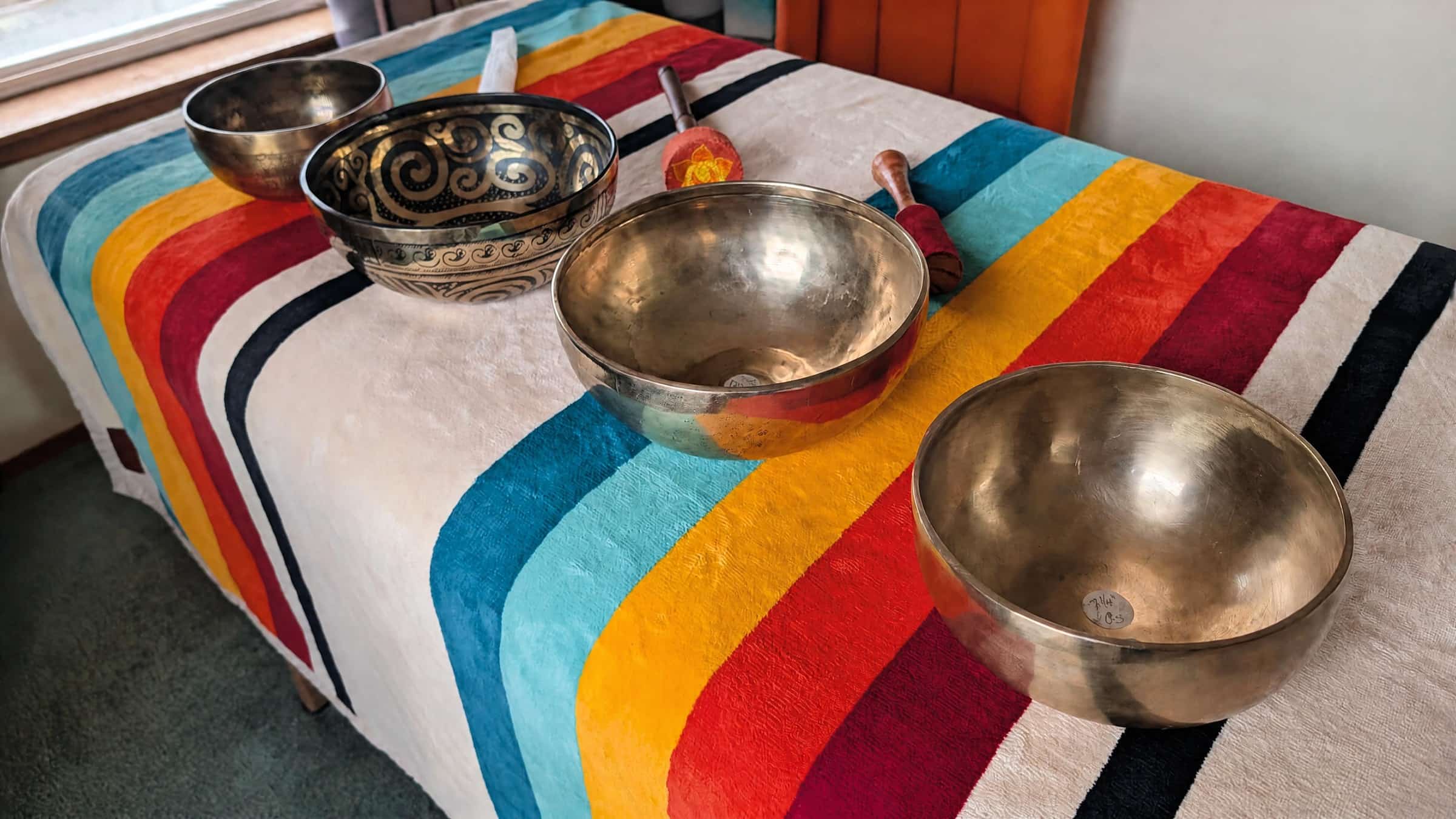 Four Tibetan singing bowls and mallets on a treatment table with a rainbow-striped blanket in Lara’s private sound healing room in Anacortes.